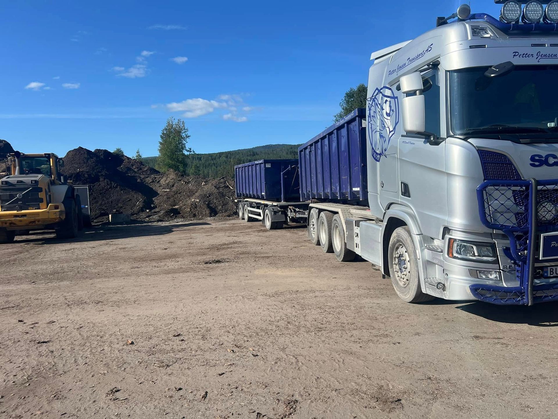 Sølvfarget semitrailer med blå containere parkert på en grusplass, med en haug med treavfall