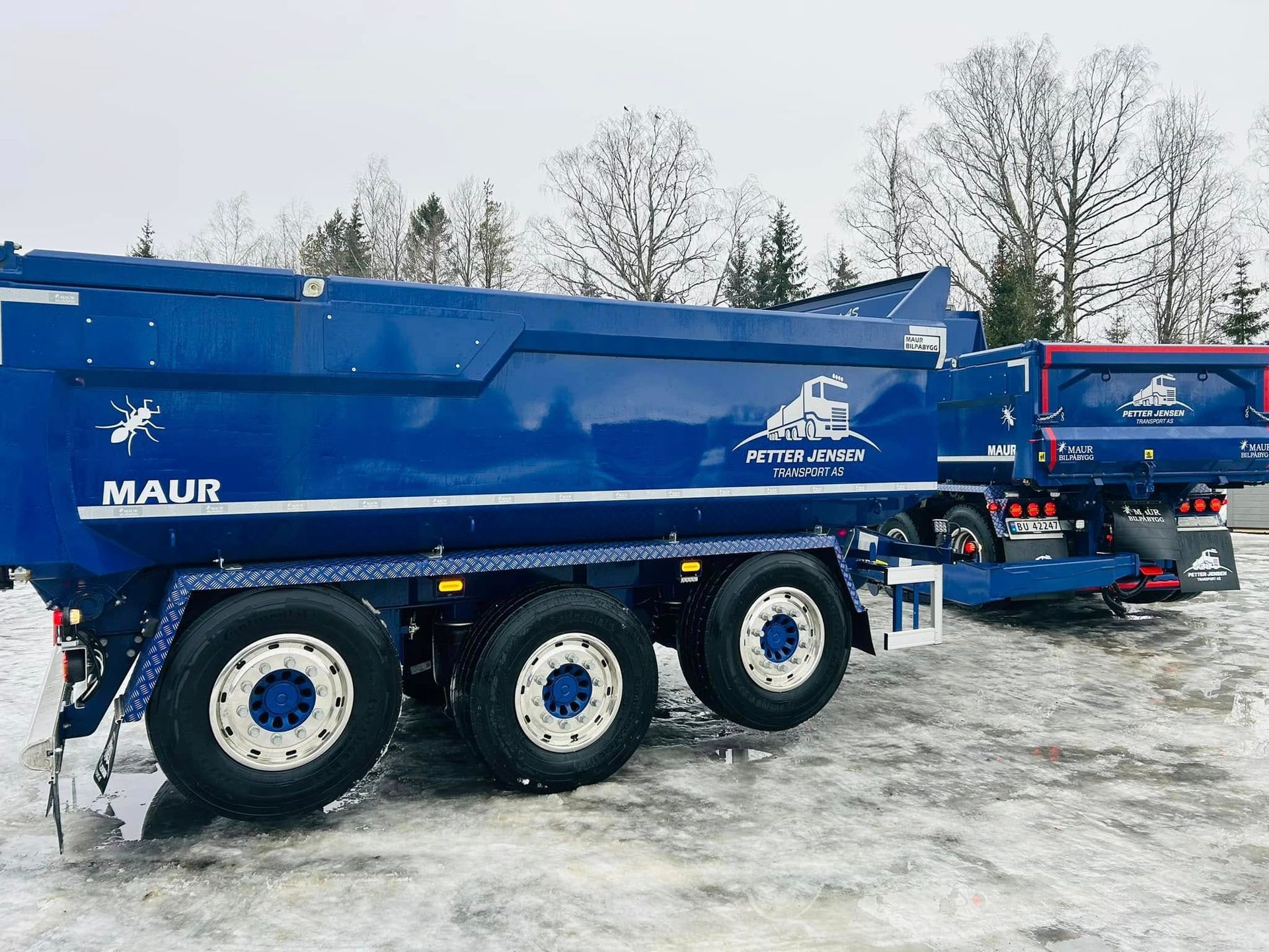 En mørkeblå tippbil med tilhenger står parkert i et snødekt utendørsområde. Den har hvite detaljer og logoer.