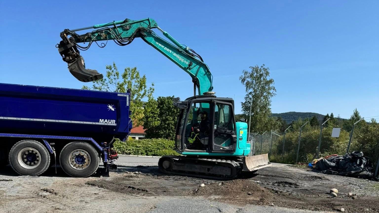 En blågrønn gravemaskin laster en blå dumper med jord. Sett mot en lys blå himmel og spredte grønne trær.