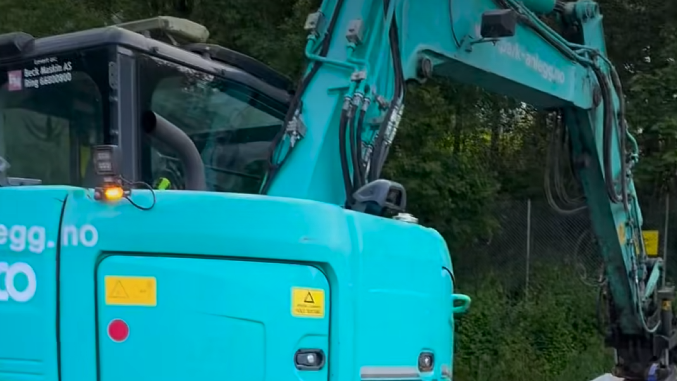 En blågrønn gravemaskin med en utstrakt arm nær grønt løvverk. Gule varselklistremerker er synlige på karosseriet.