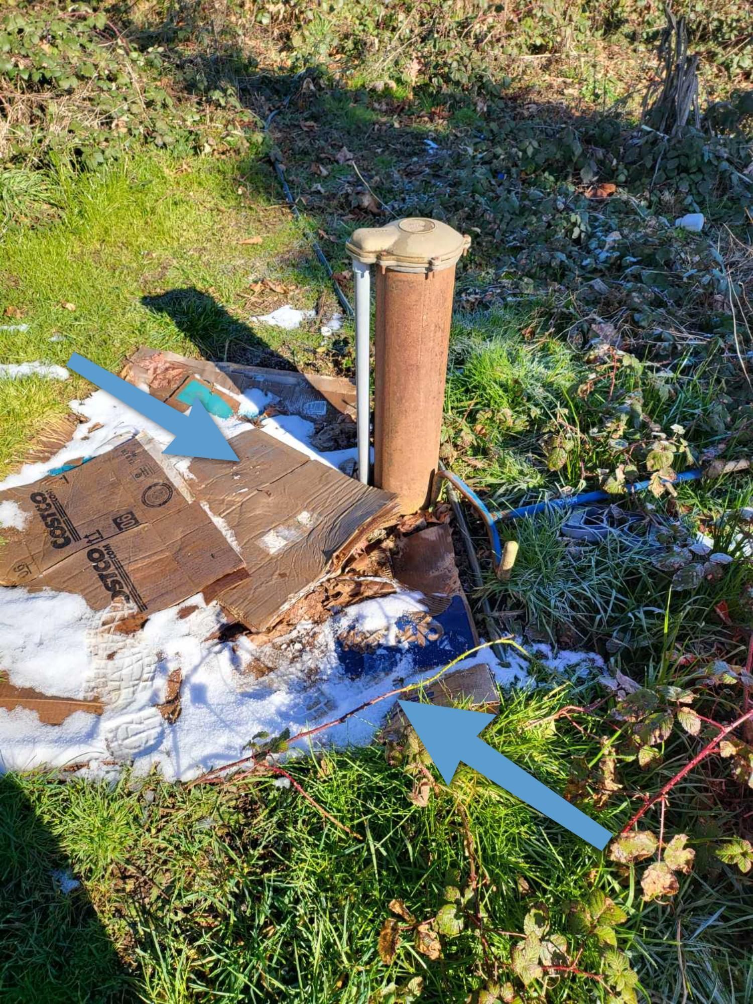 A cardboard box is sitting in the grass next to a rusty pipe.