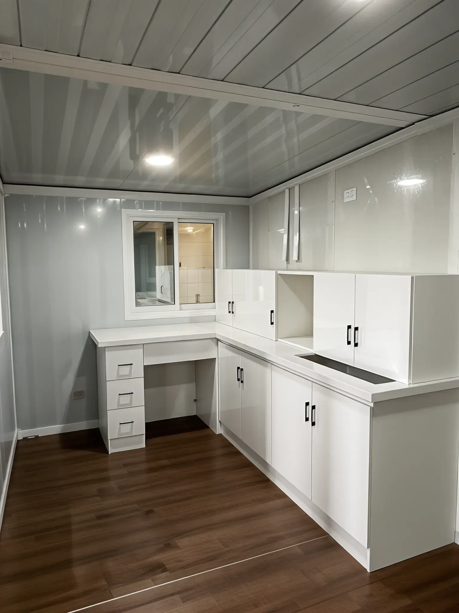 Interior of a modern kitchen with white cabinets, desk, window, and wood-look flooring. — Expandable Housing in Inala, QLD