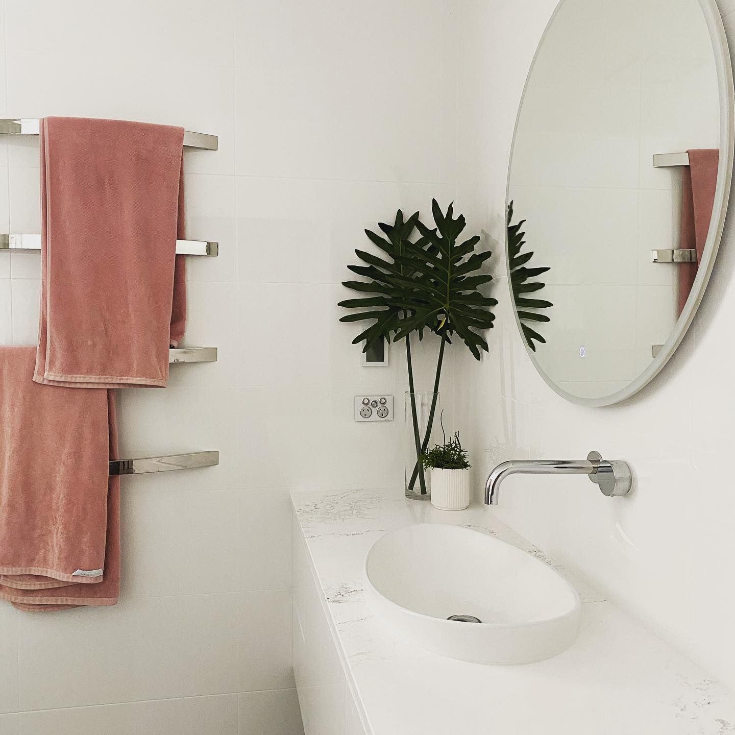 White Bathroom With Pink Towels, a Round Mirror, and a Plant Next to the Sink — Huddo's Plumbing Service in Wallalong, NSW