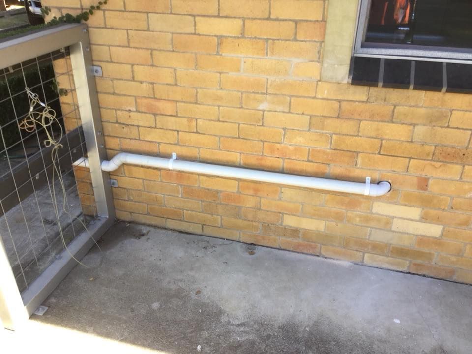White Pipe Attached to a Brick Wall, Exiting Near a Gate on a Concrete Surface — Huddo's Plumbing Service in Wallalong, NSW