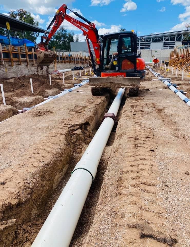 A Small Excavator Digs a Trench Around White Pipes — Huddo's Plumbing Service in Hunter Valley, NSW
