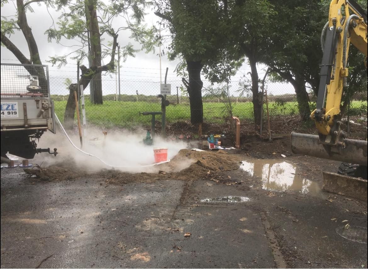An Excavator and a Truck Spewing Steam and Water on Asphalt — Huddo's Plumbing Service in Newcastle, NSW