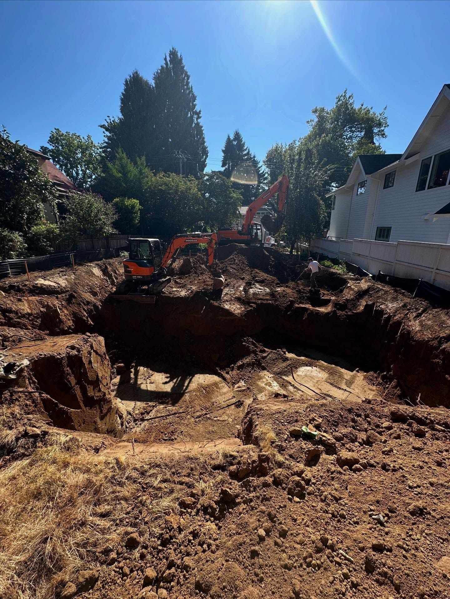 Excavator digging a large dirt pit in a sunny residential yard beside a white house.