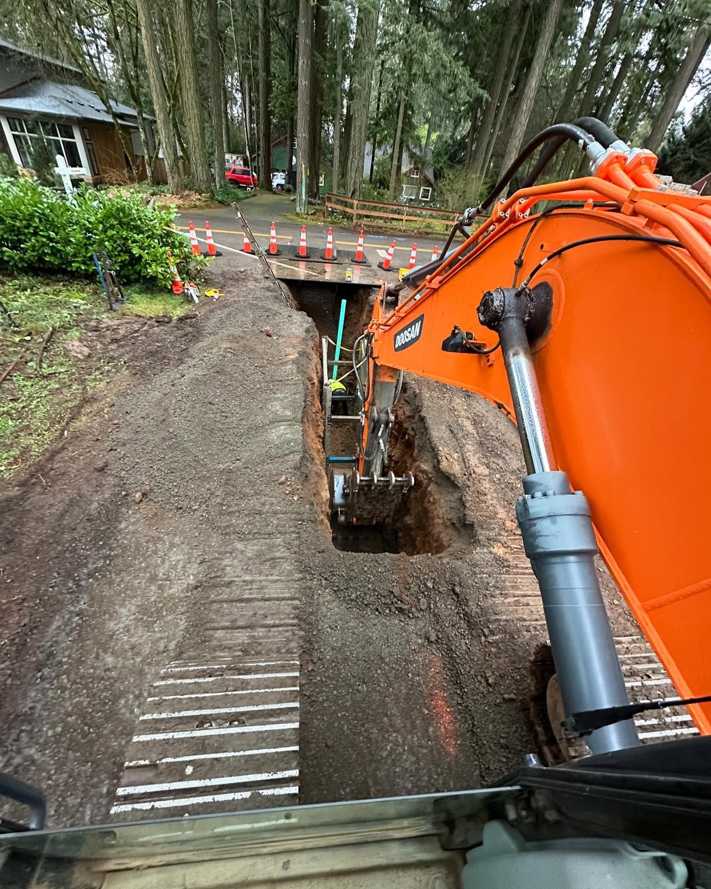 Excavator digging a trench beside a house, with a concrete culvert under a dirt driveway in a wooded area