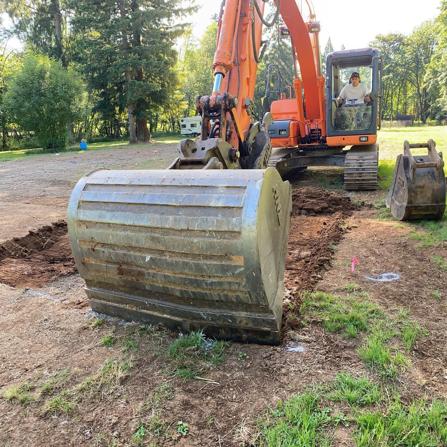 Orange excavator with a large metal bucket in a wooded dirt clearing