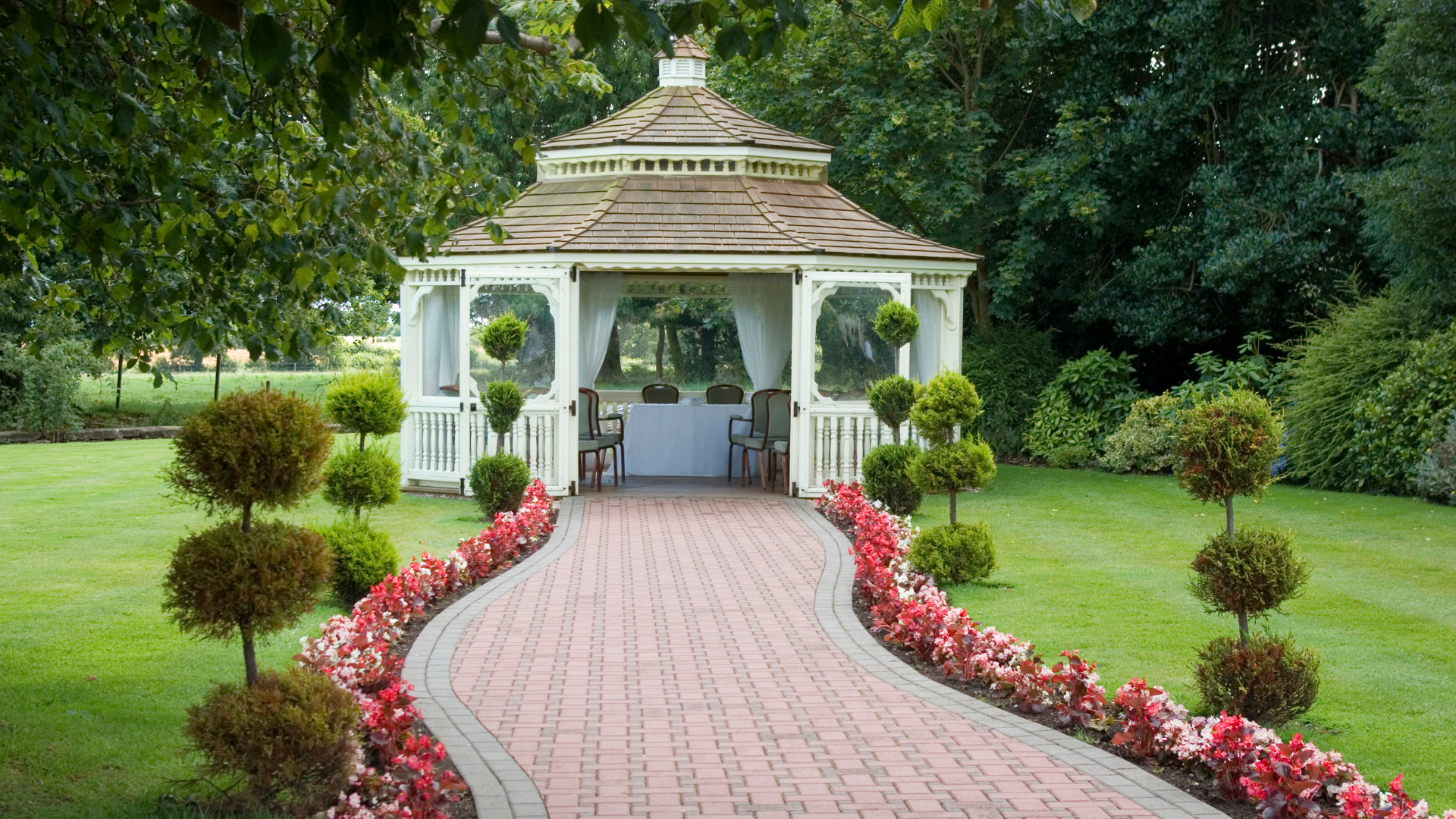 A white gazebo is surrounded by flowers and trees in a park.