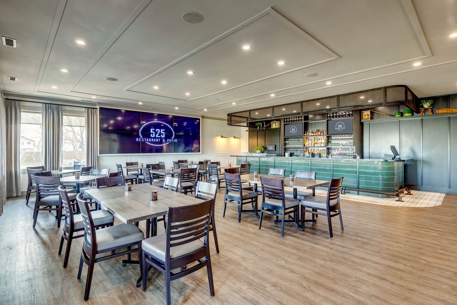 A spacious dining area featuring wooden tables, gray upholstered chairs, a long green bar, and a large wall display.
