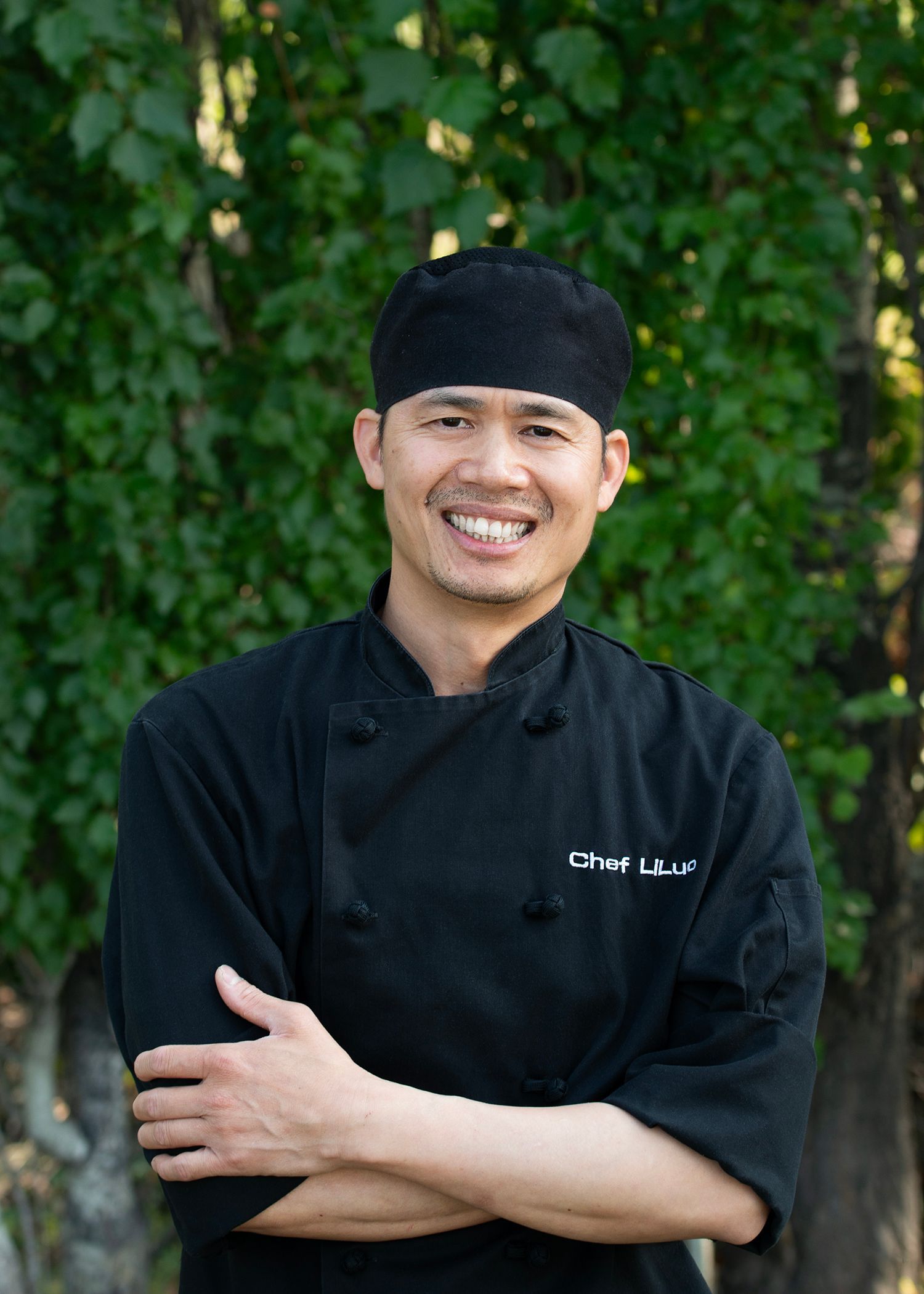 A smiling chef in a black uniform and skull cap with arms crossed, standing in front of green foliage.