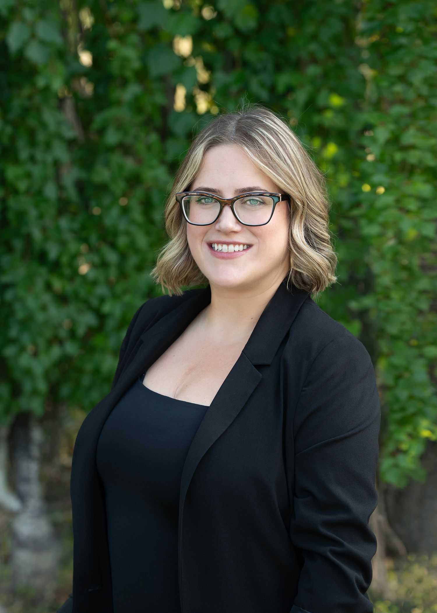 A smiling professional wearing a black blazer and glasses, standing outdoors in front of a green leafy backdrop.