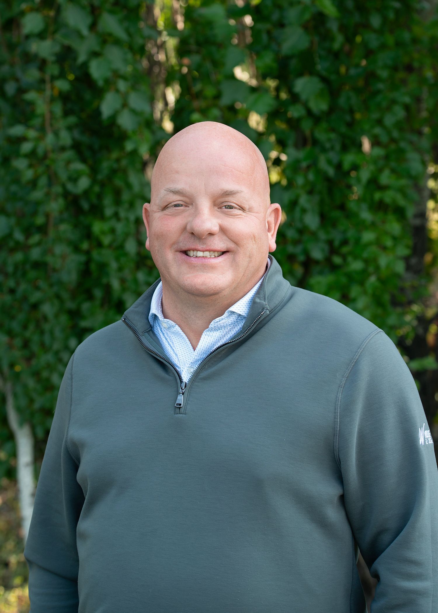 Man with short blonde hair, smiling, in a white patterned shirt, standing in front of a brick wall.