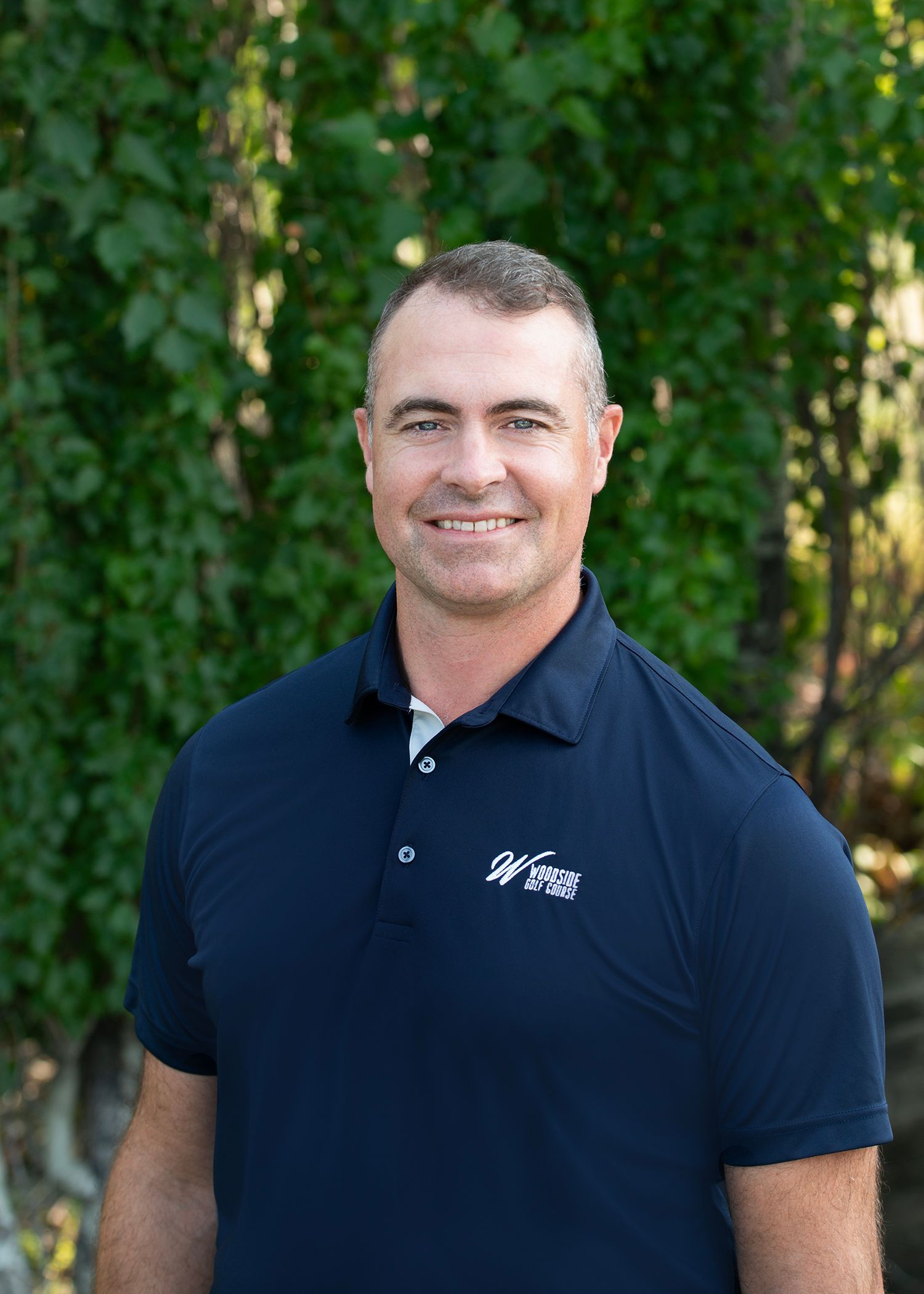 A man with short hair wearing a navy blue polo shirt with a logo, standing in front of green foliage.