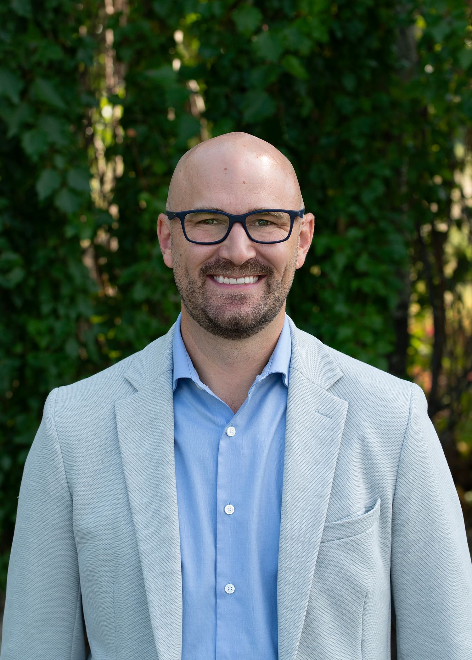 Man with short blonde hair, smiling, in a white patterned shirt, standing in front of a brick wall.