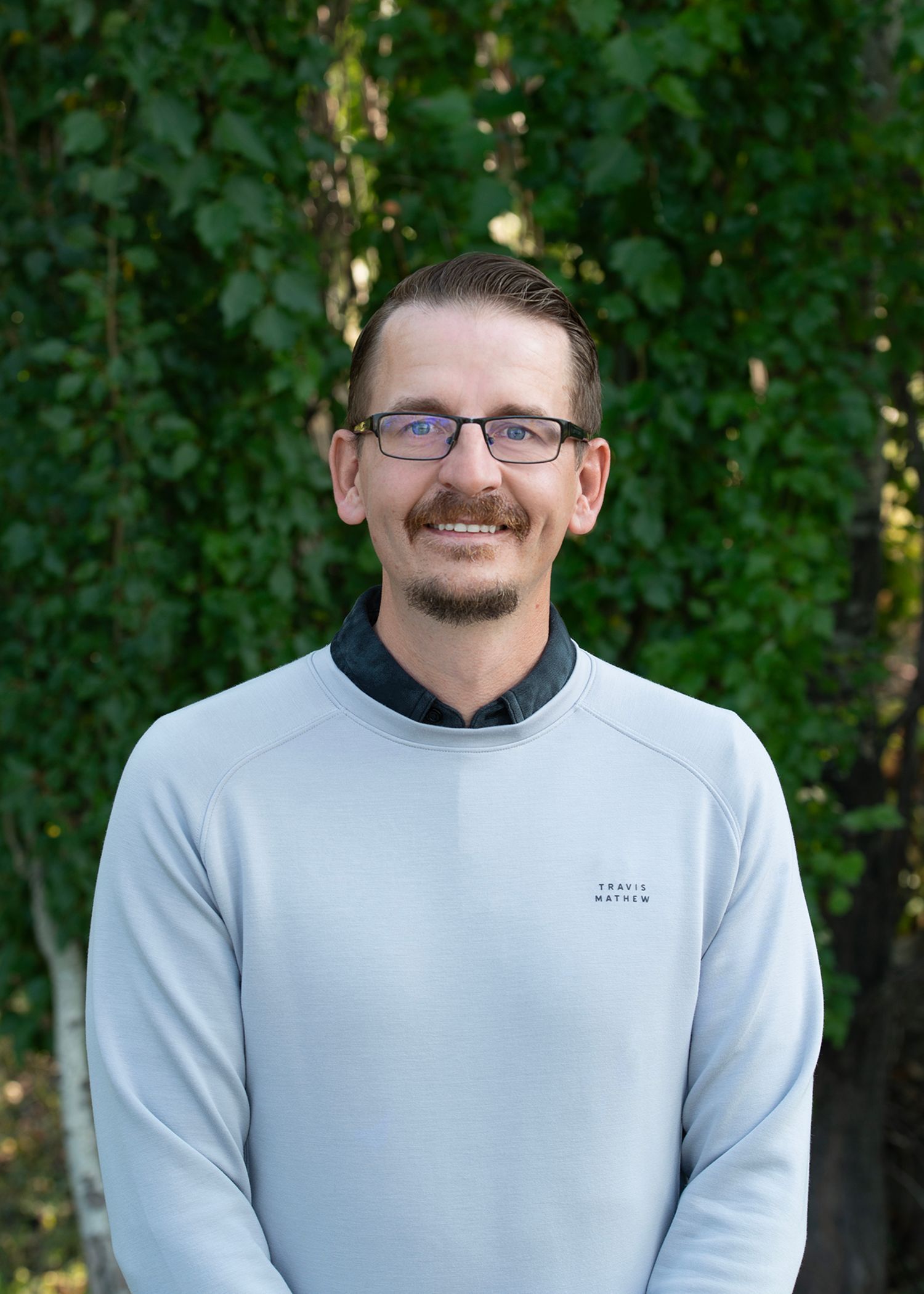 A man wearing glasses, blue sweater, and gray pants stands in front of a brick wall, with a golf course in the background.