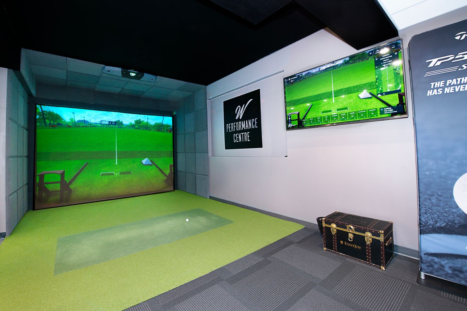 Indoor golf simulator room with a green turf mat, large digital screen, and a storage trunk on a gray carpeted floor.