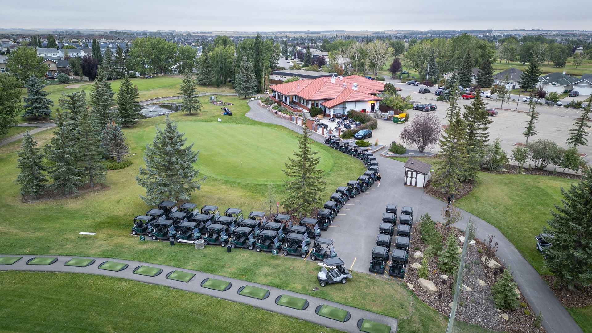 An aerial view of a golf course with golf carts lined up, near a clubhouse, with trees and a cloudy sky.