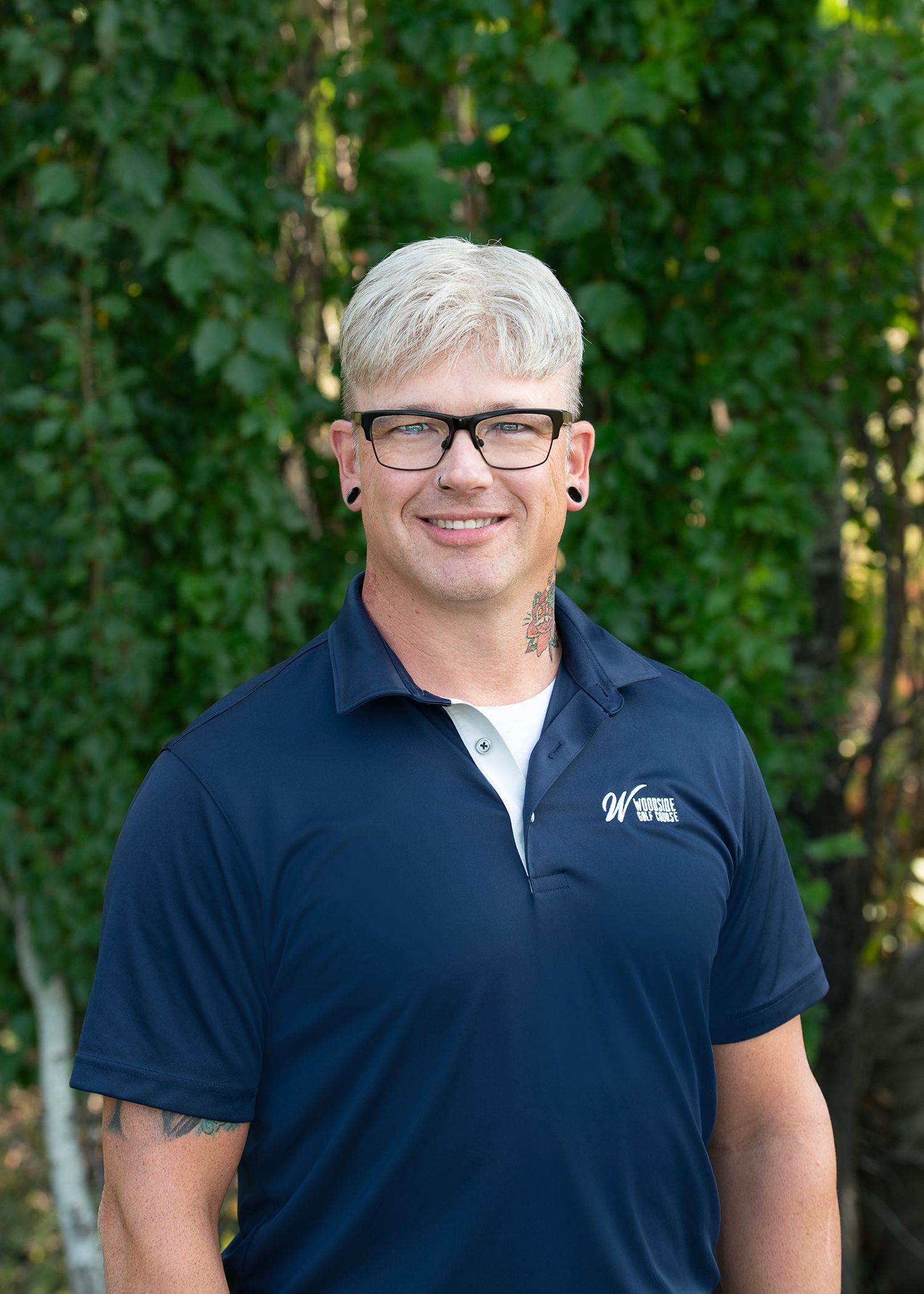 Person with light hair and glasses wearing a blue polo shirt, smiling, in front of a green leafy background.