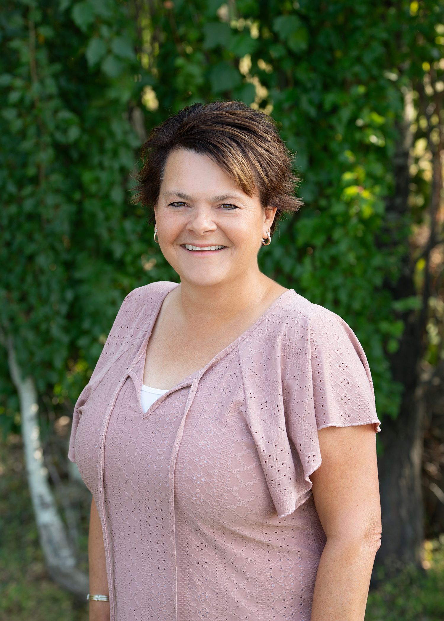 A smiling person wearing a textured, mauve-colored short-sleeve top, positioned outdoors in front of a leafy green tree.