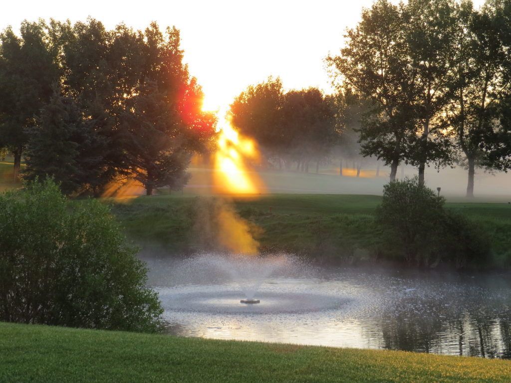 Sunrise over a misty pond with a fountain, green grass, and trees. Bright sunlight shines through.