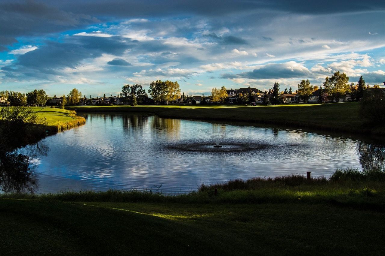 Lake in a park setting with a fountain. Trees and houses line the horizon under a cloudy sky.