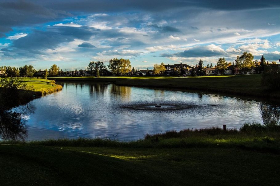 Lake in a park setting with a fountain. Trees and houses line the horizon under a cloudy sky.