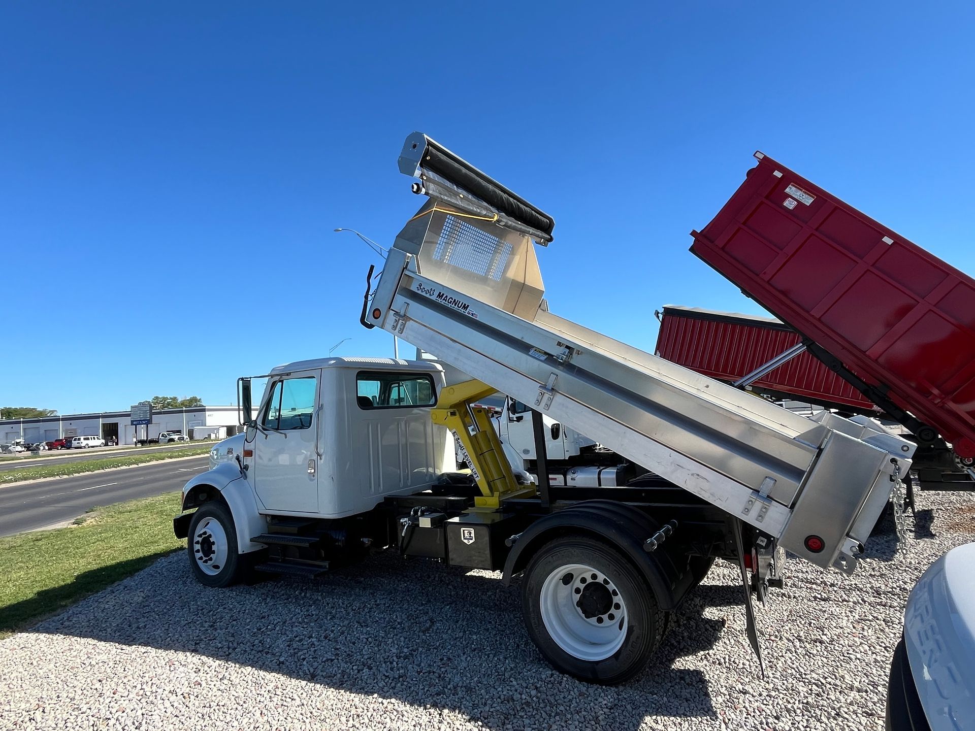 A white dump truck is parked on gravel next to a red dump truck.