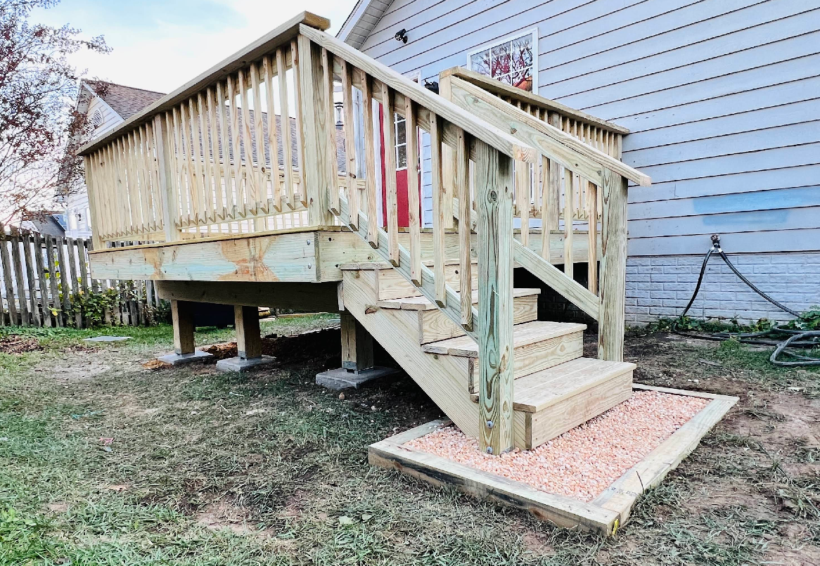 Wooden deck with stairs leading down to a gravel landing; built next to a house with white siding and a red door.
