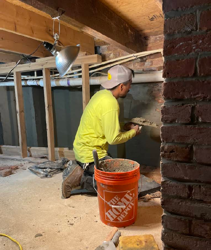 A worker in a yellow shirt kneels, applying mortar to a wall next to an orange bucket.