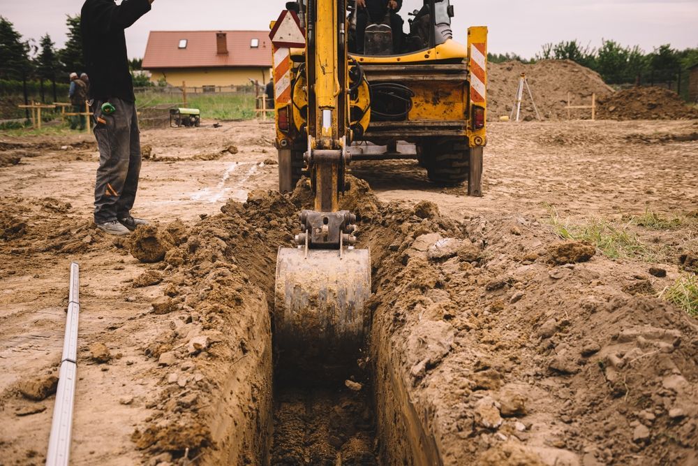 Yellow excavator digging a trench in dirt, worker standing nearby a home under construction.