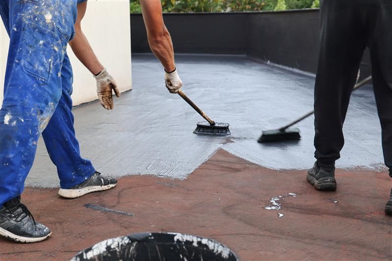 Three people applying a black sealant to a rooftop with brushes, wearing work clothes and gloves.