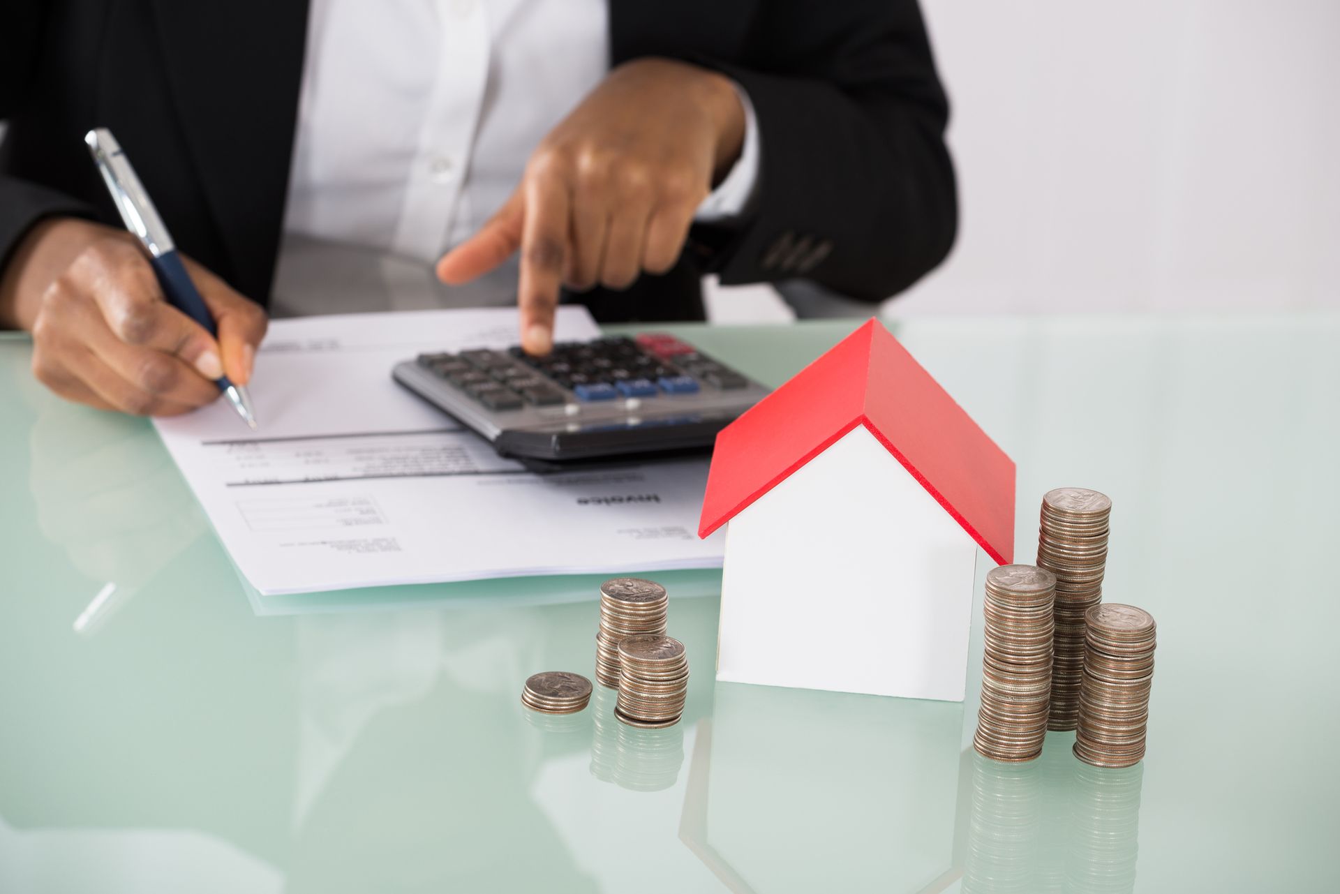 A woman is sitting at a desk using a calculator next to a model house and stacks of coins.