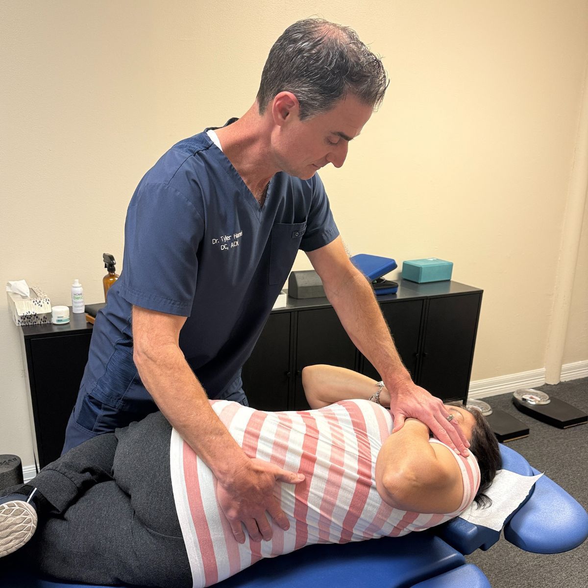 Chiropractor adjusting patient's spine on a massage table. The man is wearing blue scrubs.