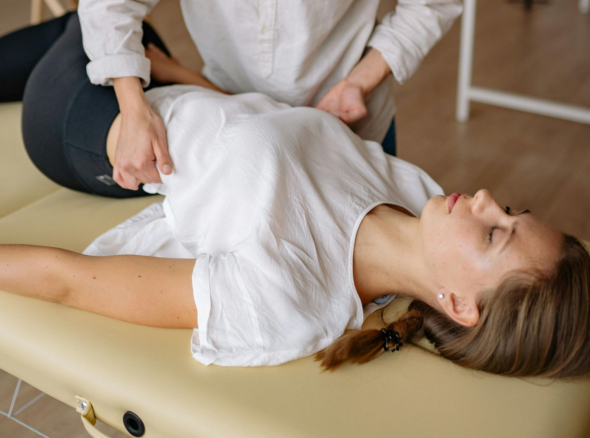 Woman receiving a hip adjustment on a massage table. Hands on hip, white shirt, indoor setting.