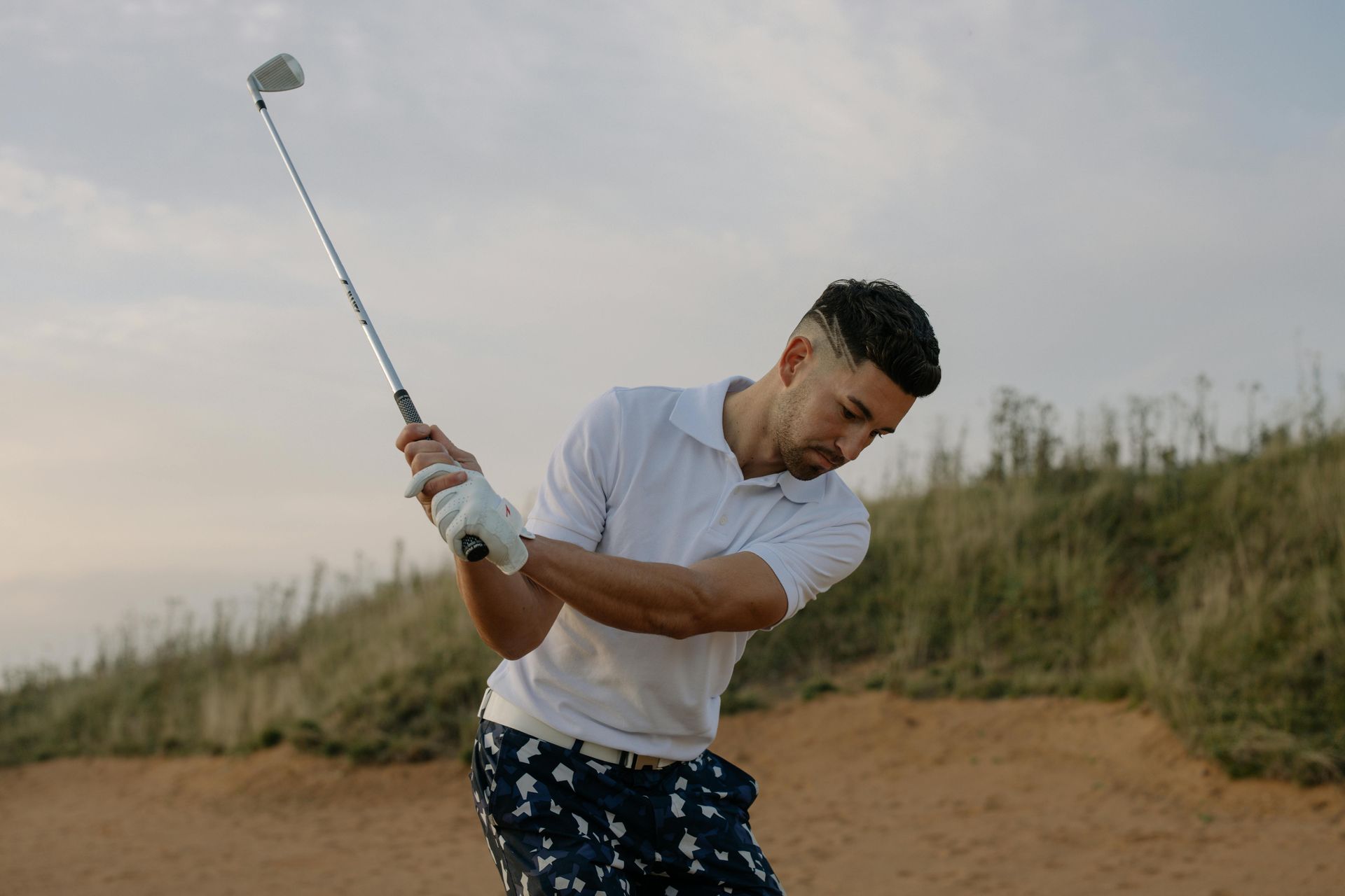 Golfer swings club on a sand bunker, wearing a white polo shirt and patterned shorts.