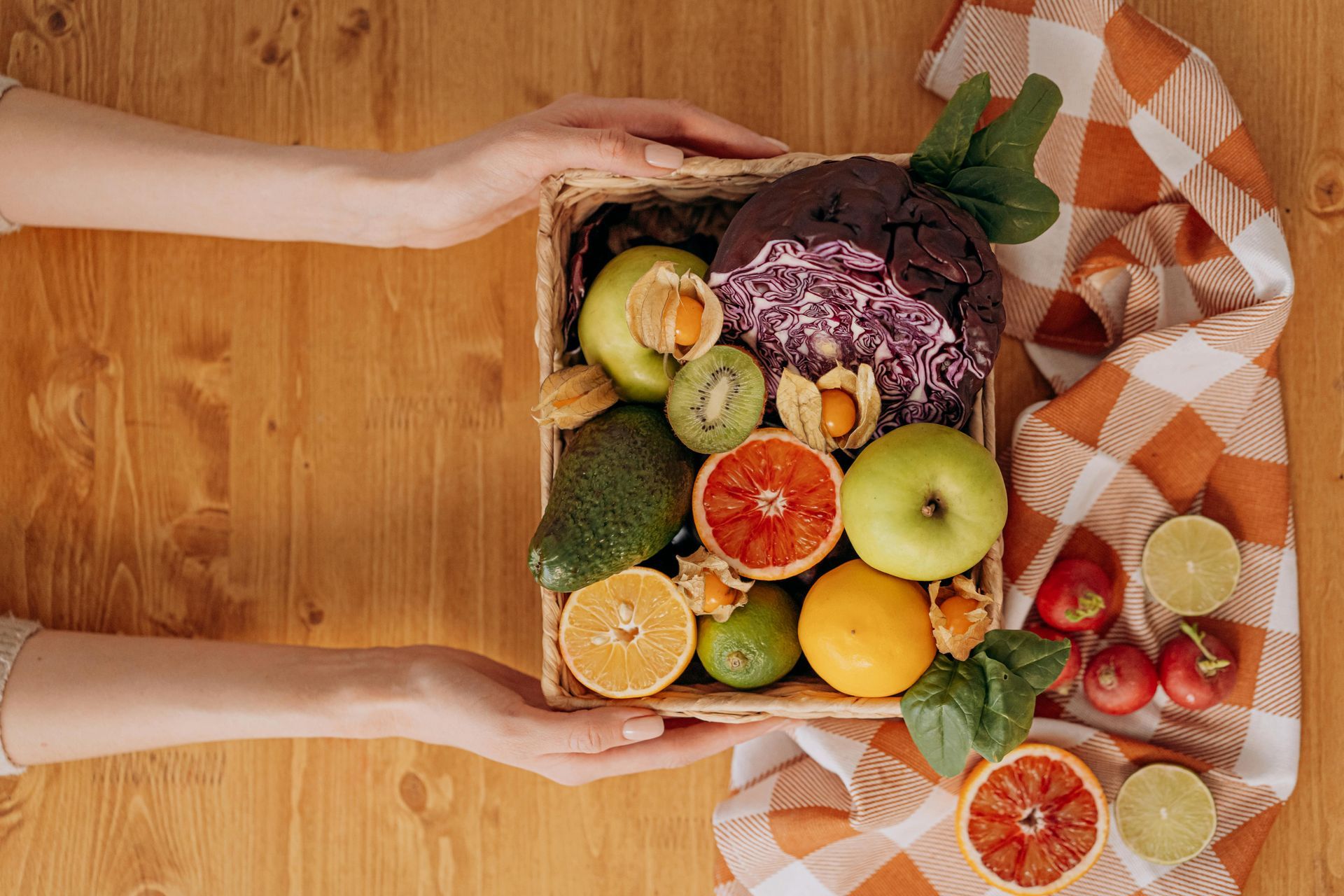 Hands holding a basket filled with colorful fruits and vegetables on a wooden table, next to a checkered cloth.