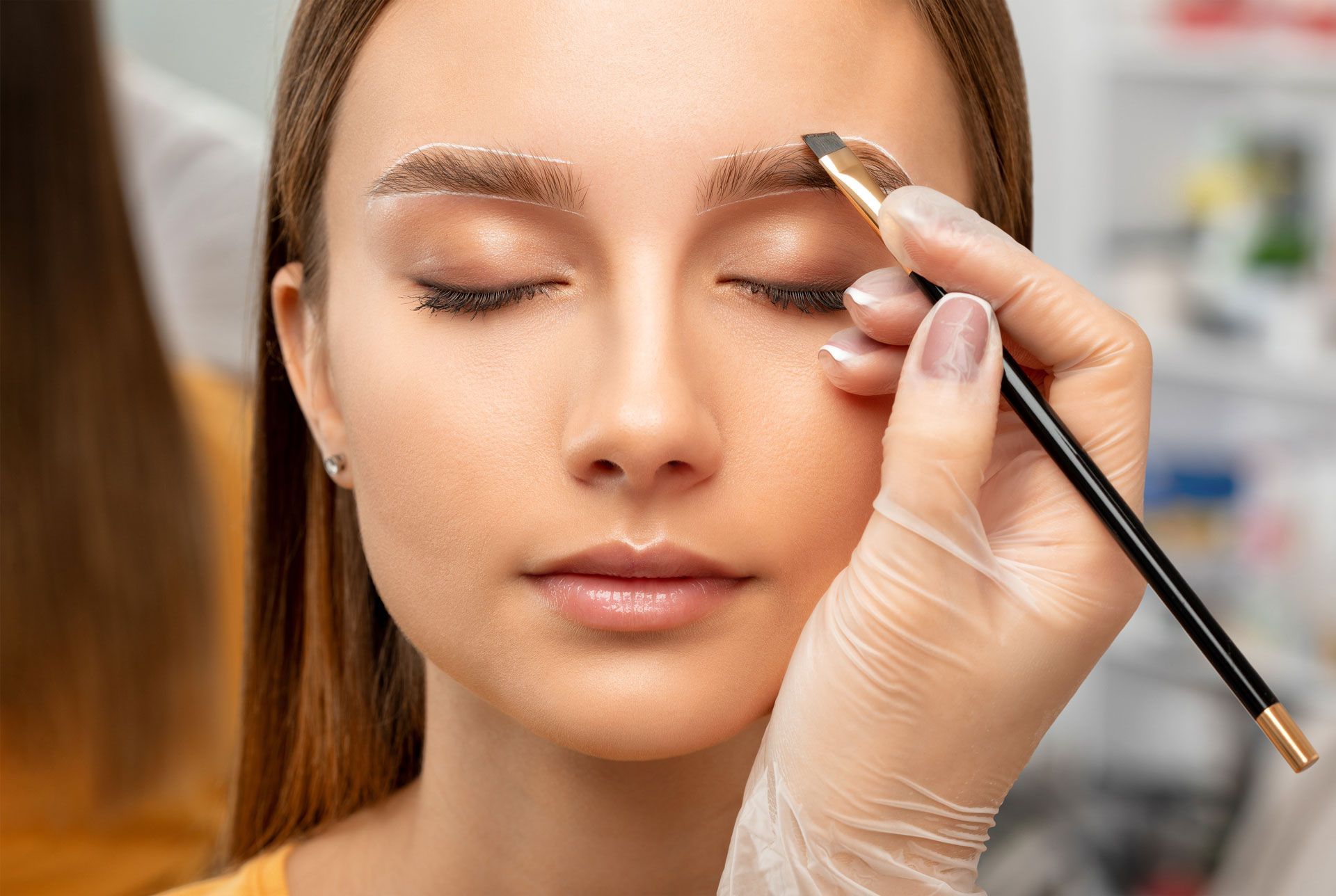 A person having their eyebrow shaped with a brush, close-up. Light skin, eyes closed. White room, gloved hand.