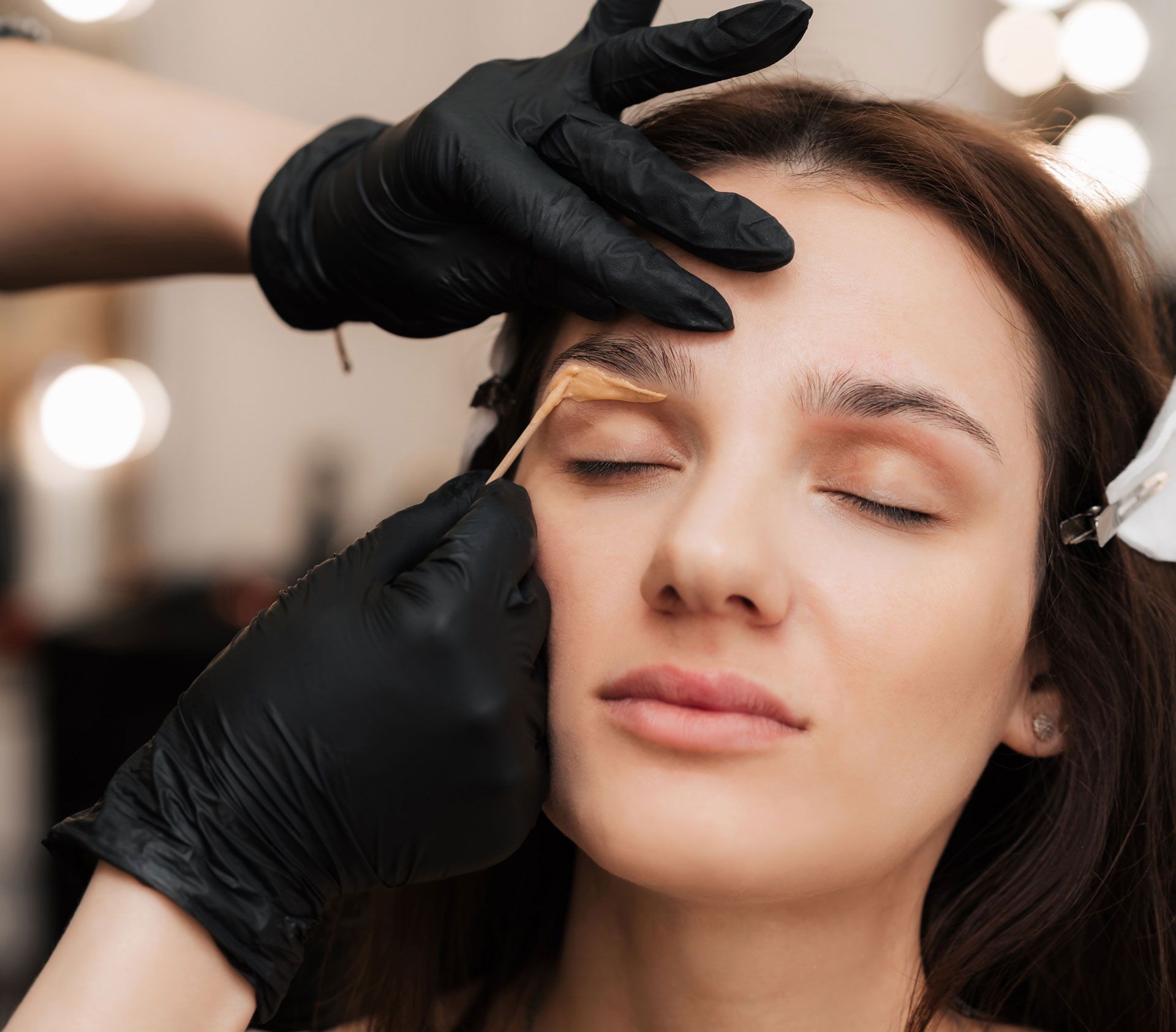 Woman having eyebrow treatment; black-gloved hands applying product. Indoors, eyes closed.