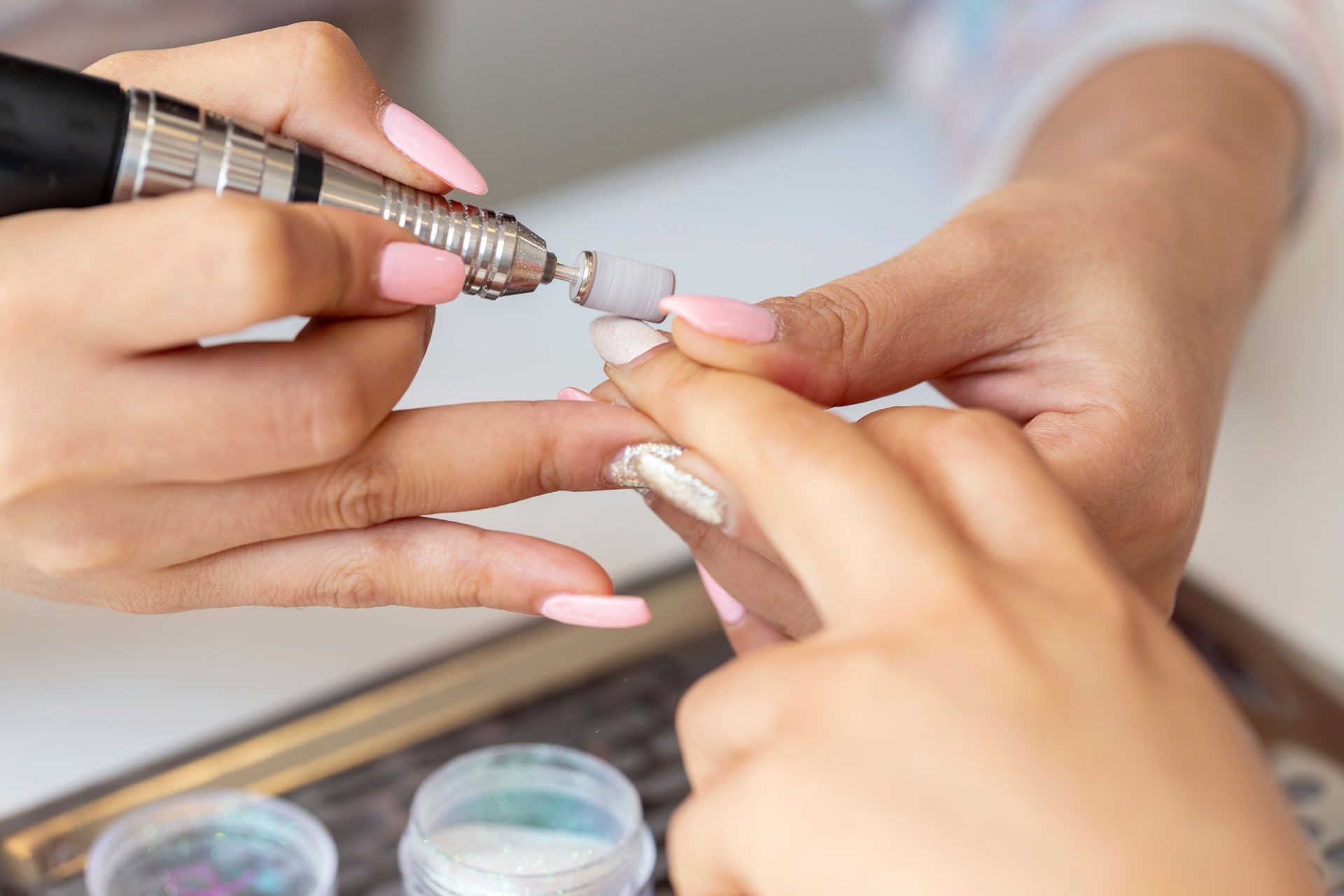 Manicurist using a nail drill on a client's pink acrylic nail. White table, jars of powder are visible.