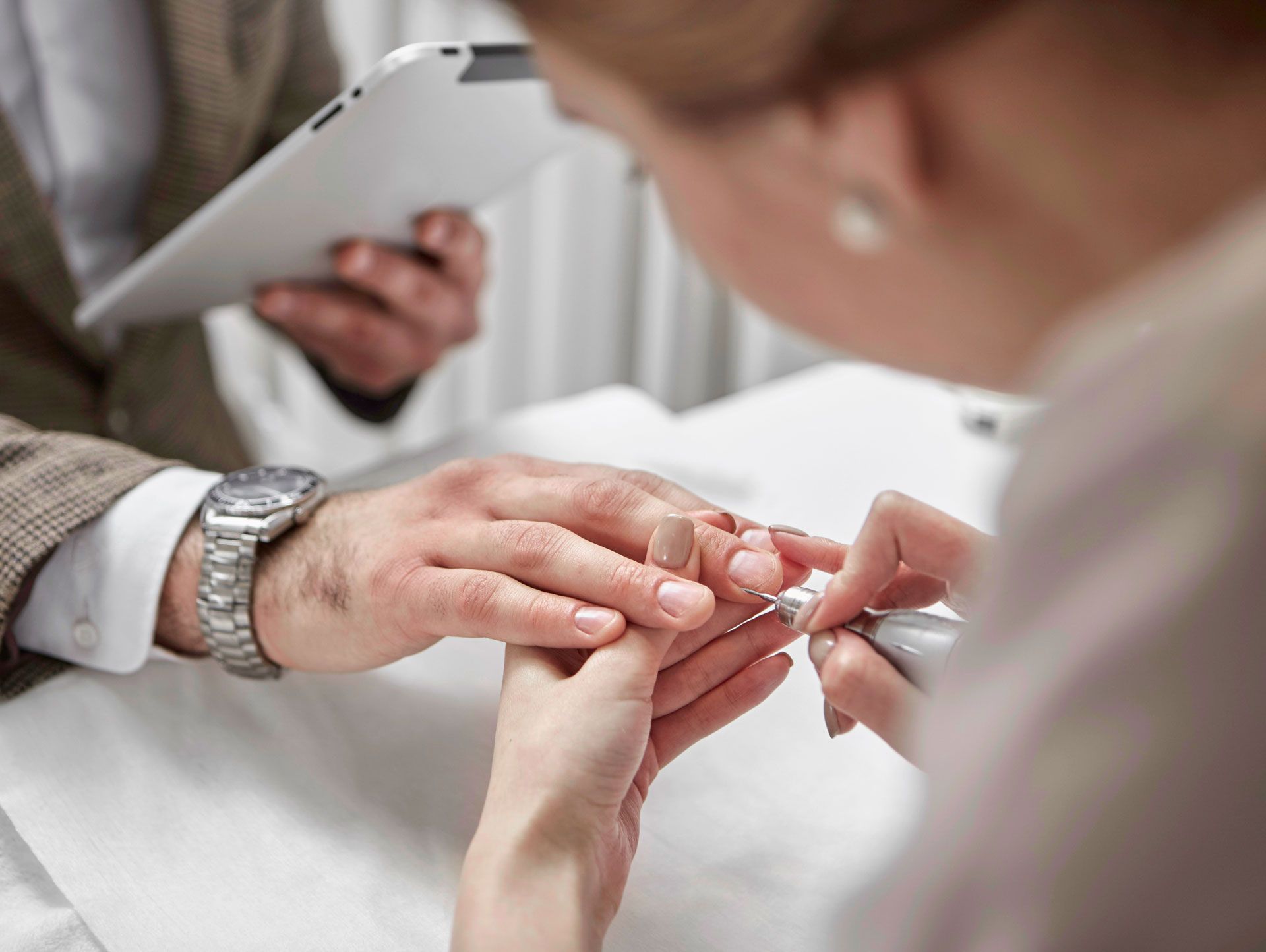Man receiving manicure, technician using a tool on his nails. Person holding tablet in background.