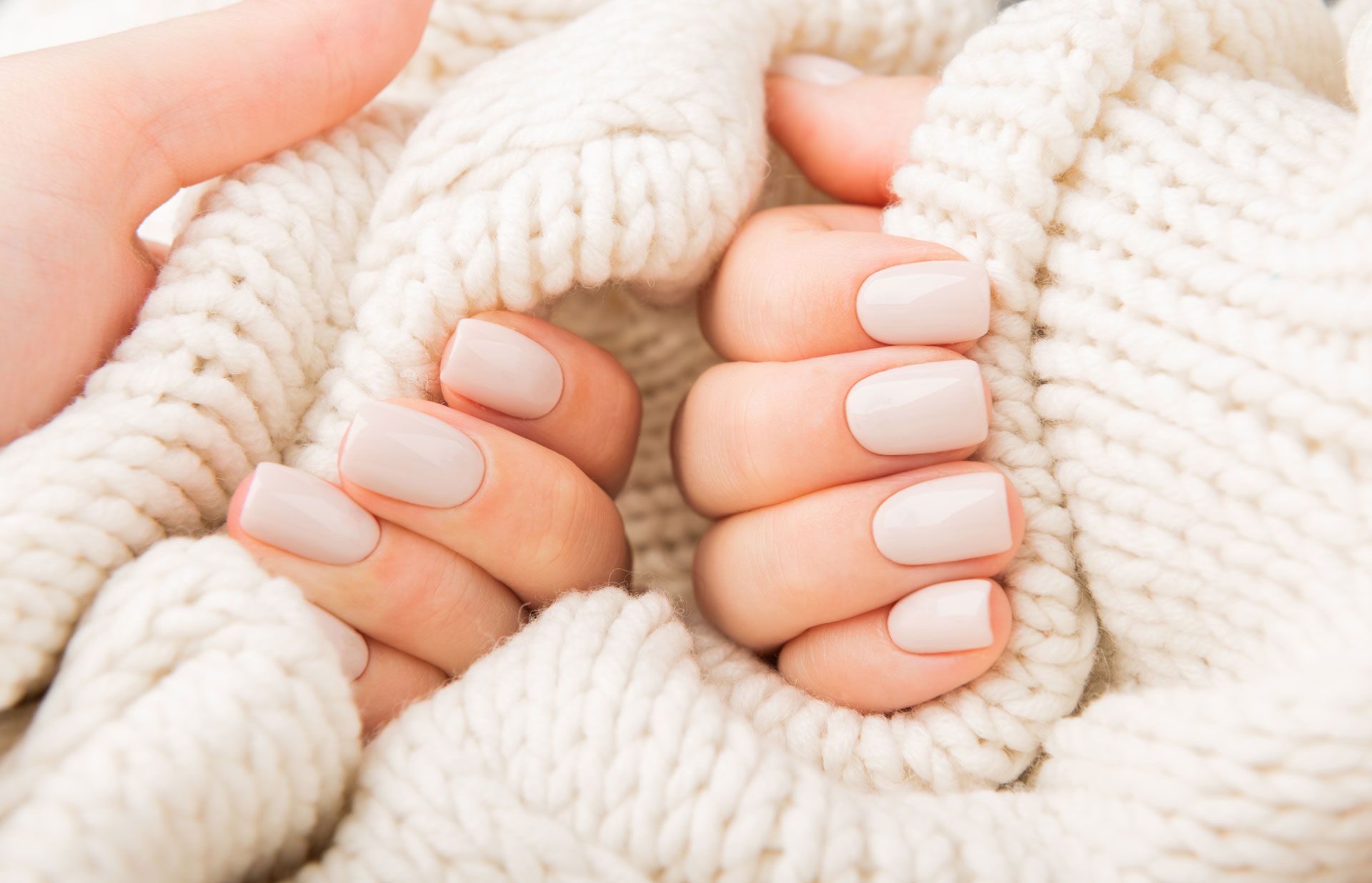 Hands with light pink nails wrapped in a thick, cream-colored knitted sweater.