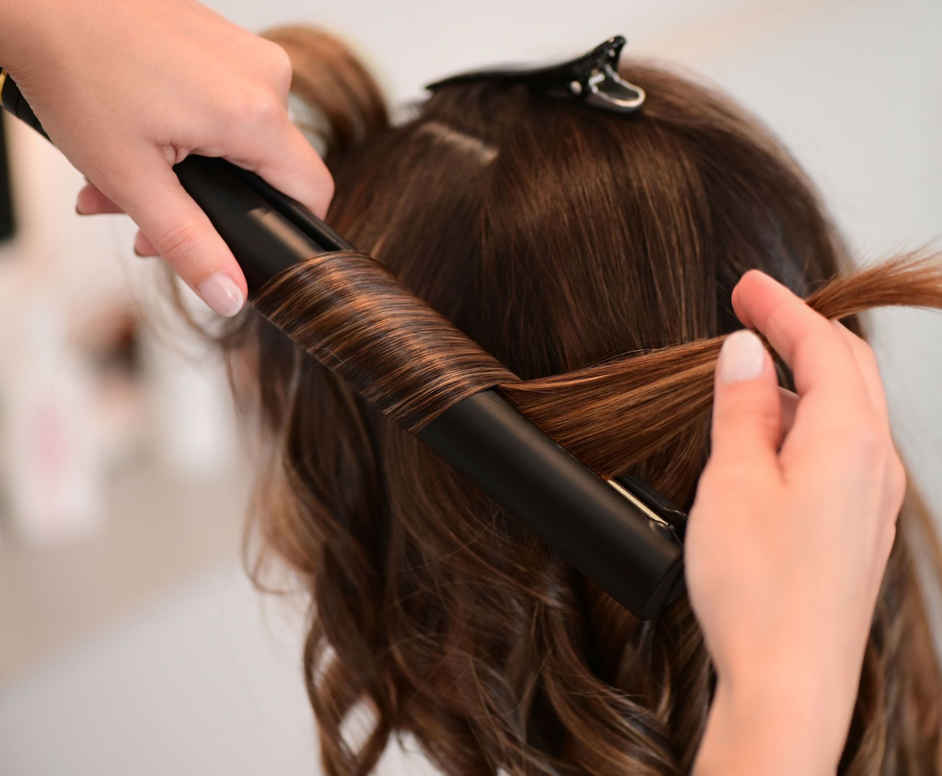 Hands curling brown hair with a curling iron in a salon setting.
