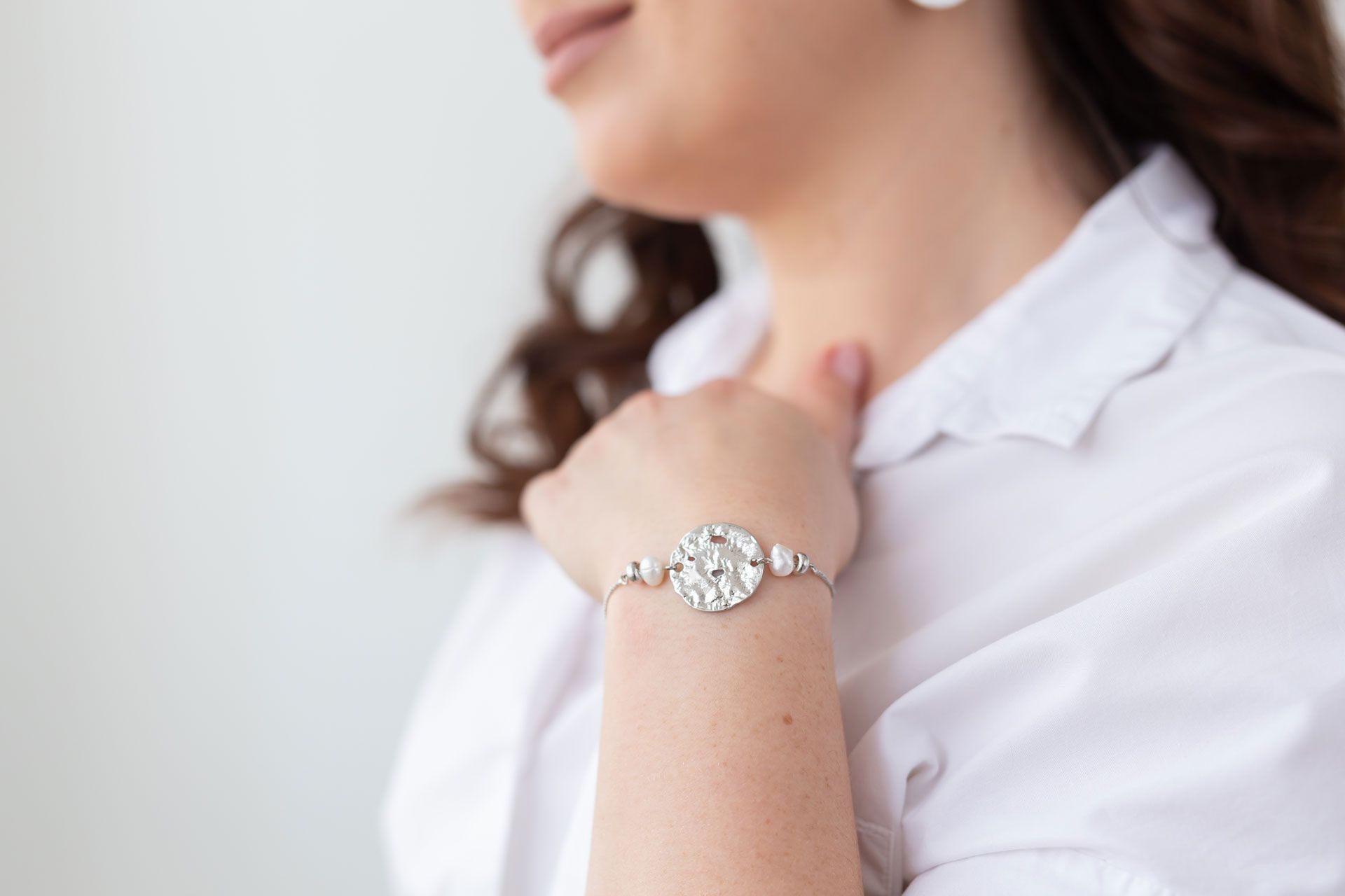 Woman wearing a silver bracelet, touching her neck, with a white shirt against a white wall.