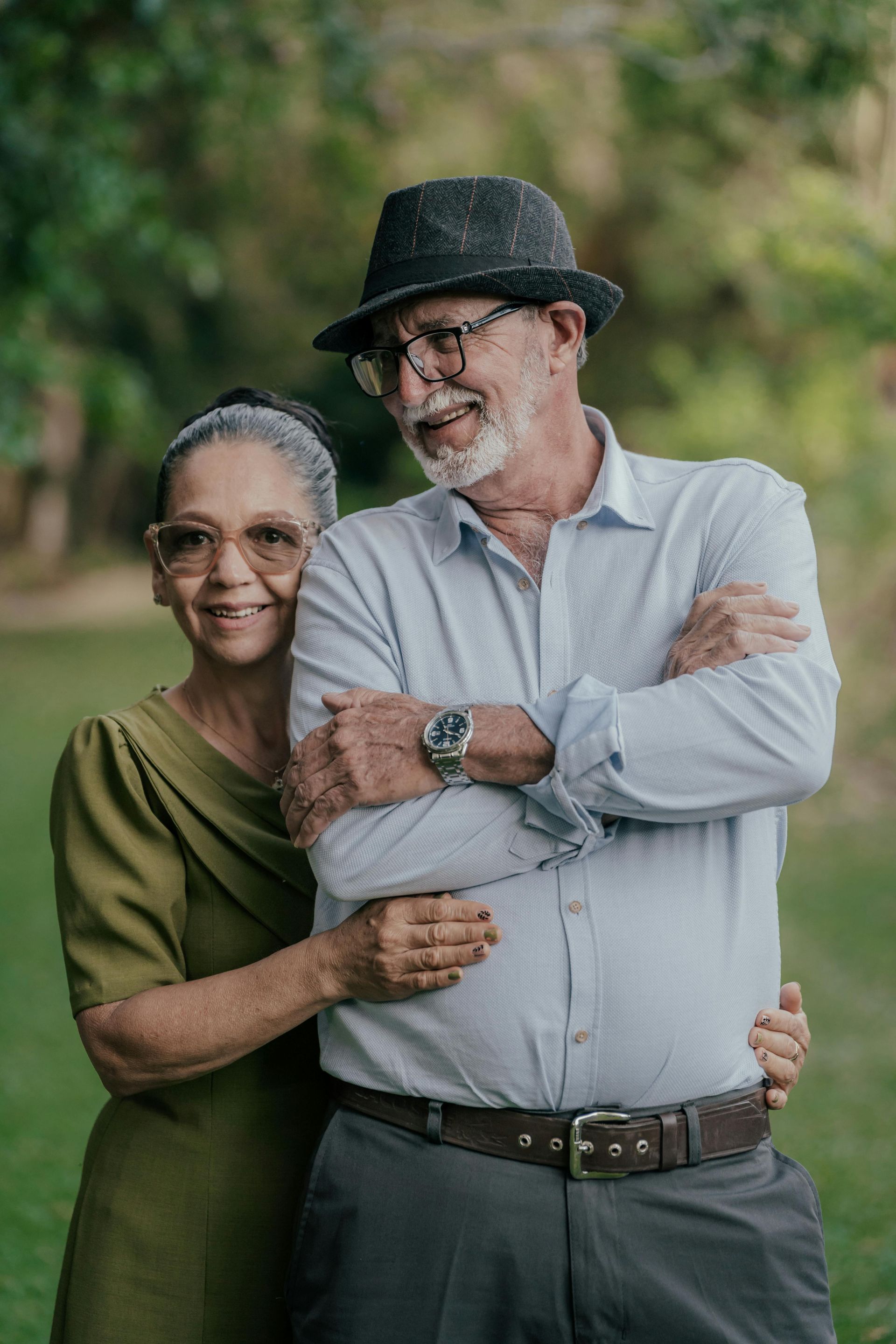 An elderly couple standing next to each other in a park with their arms crossed.