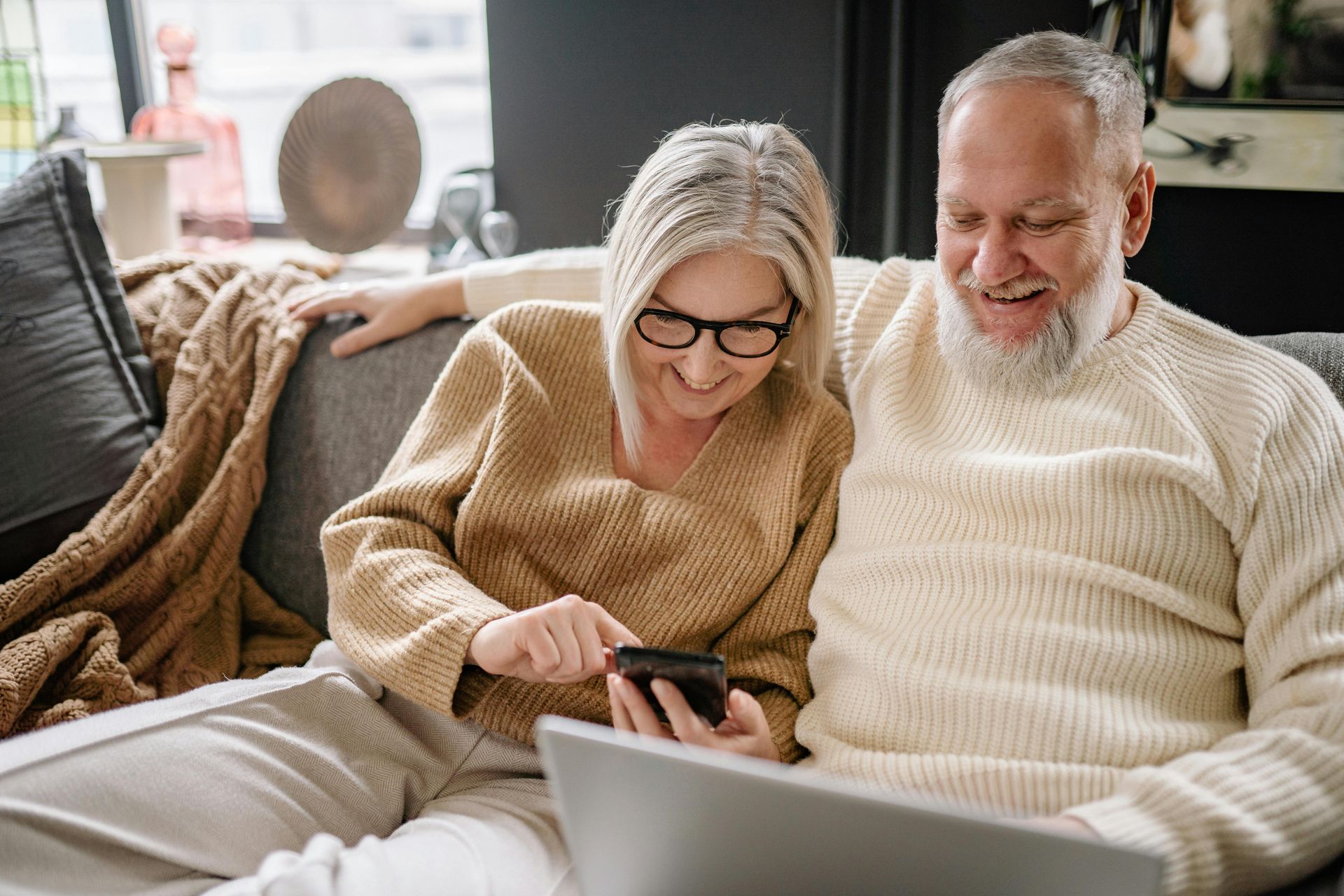 An elderly couple is sitting on a couch looking at a laptop and a cell phone.