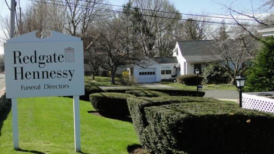 Sign for Redgate Hennessy Funeral Directors in front of a building with hedges and a grassy lawn.