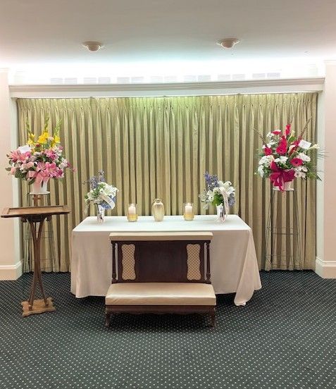 Funeral service altar with flowers, candles, and a bench, against a draped backdrop.
