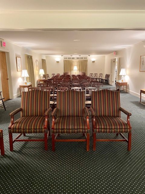 Three upholstered armchairs face rows of wooden chairs in a carpeted room with lamps.